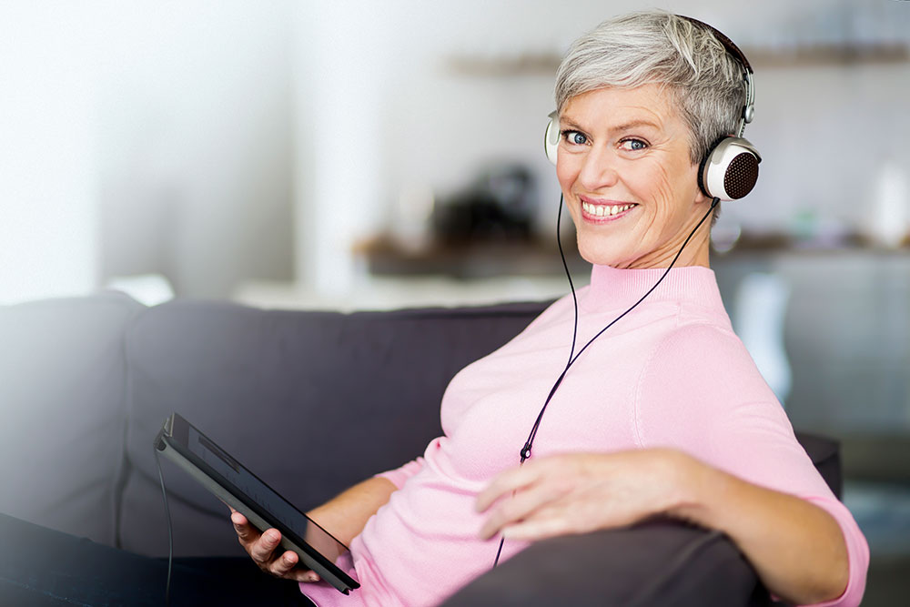 mature woman sitting on couch with digital tablet listening music with headphones