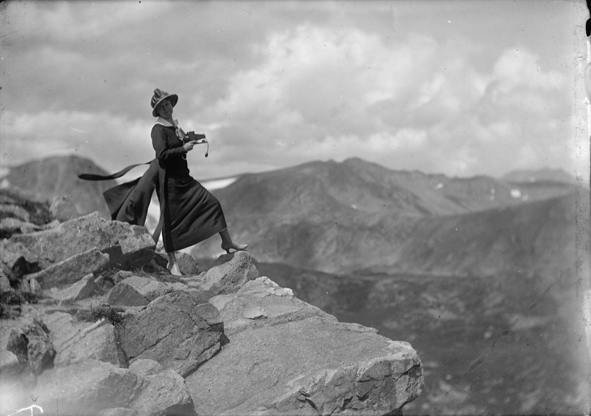 A black and white photo of a woman in a black dress standing on a mountain cliff in the mountains