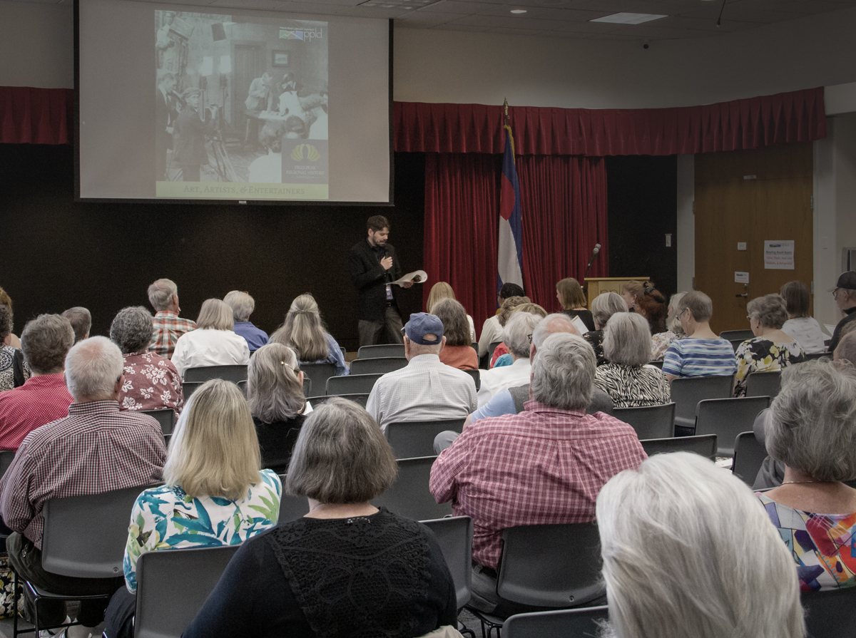 Photo of the History 2024 History Symposium. A crowd of people watch a speaker present with a screenbehind him