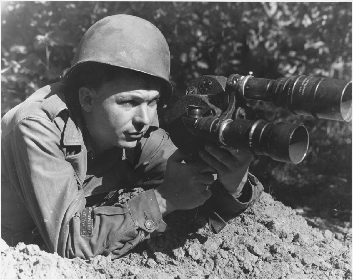An Old black and white photo of a soldier sitting on the ground prone with a big camera