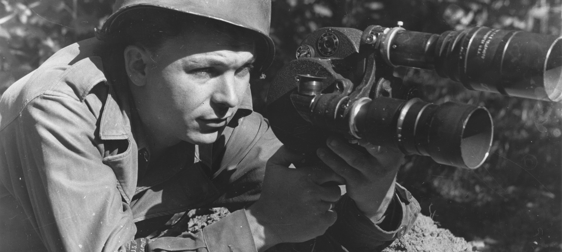 A Black and White photo of a soldier sitting prone on the ground with a big camera