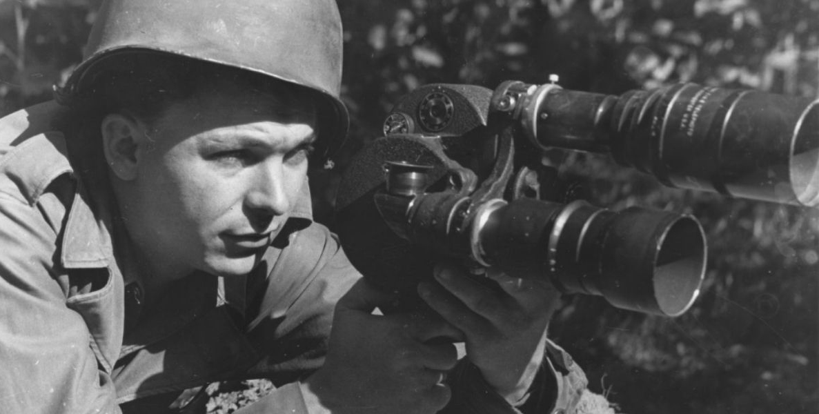 Black and White photo of a man crouched on the ground holding a large camera