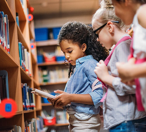 Three girls looking at a book in the library next to the shelf