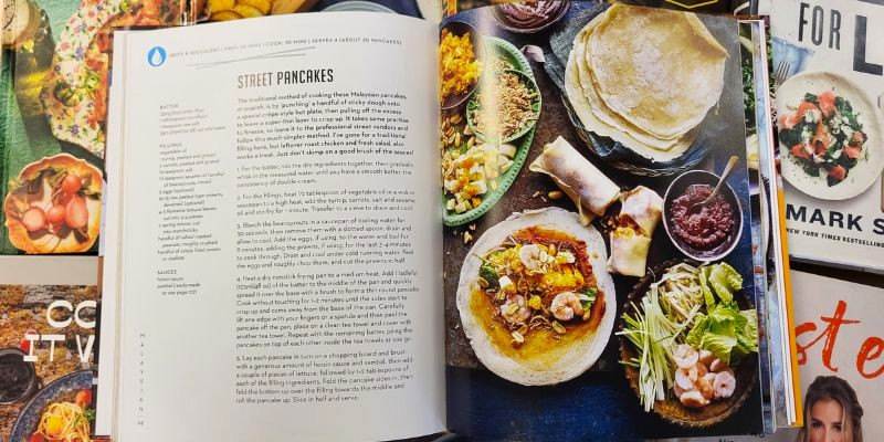 An open cookbook showing a recipe for street pancakes sits on top of a pile of other cookbooks.