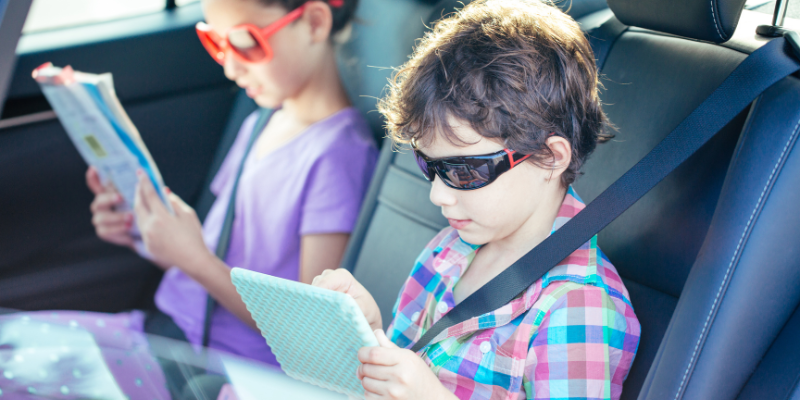 Children sit in the back seat of a vehicle, one reading while the other looks at something on a tablet. 