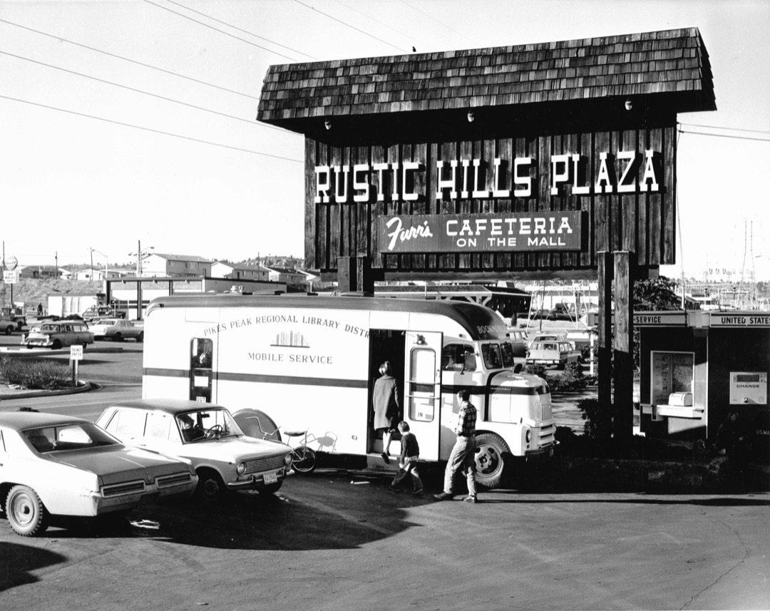 an old black and white photo of the Bookmobile parked in a parking lot in front of hte Rustic Hills Plaza in Colorado Springs surrounded by 1960s era vehicles