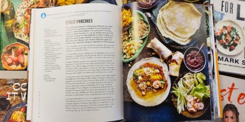 An open cookbook showing a recipe for street pancakes sits on top of a pile of other cookbooks.